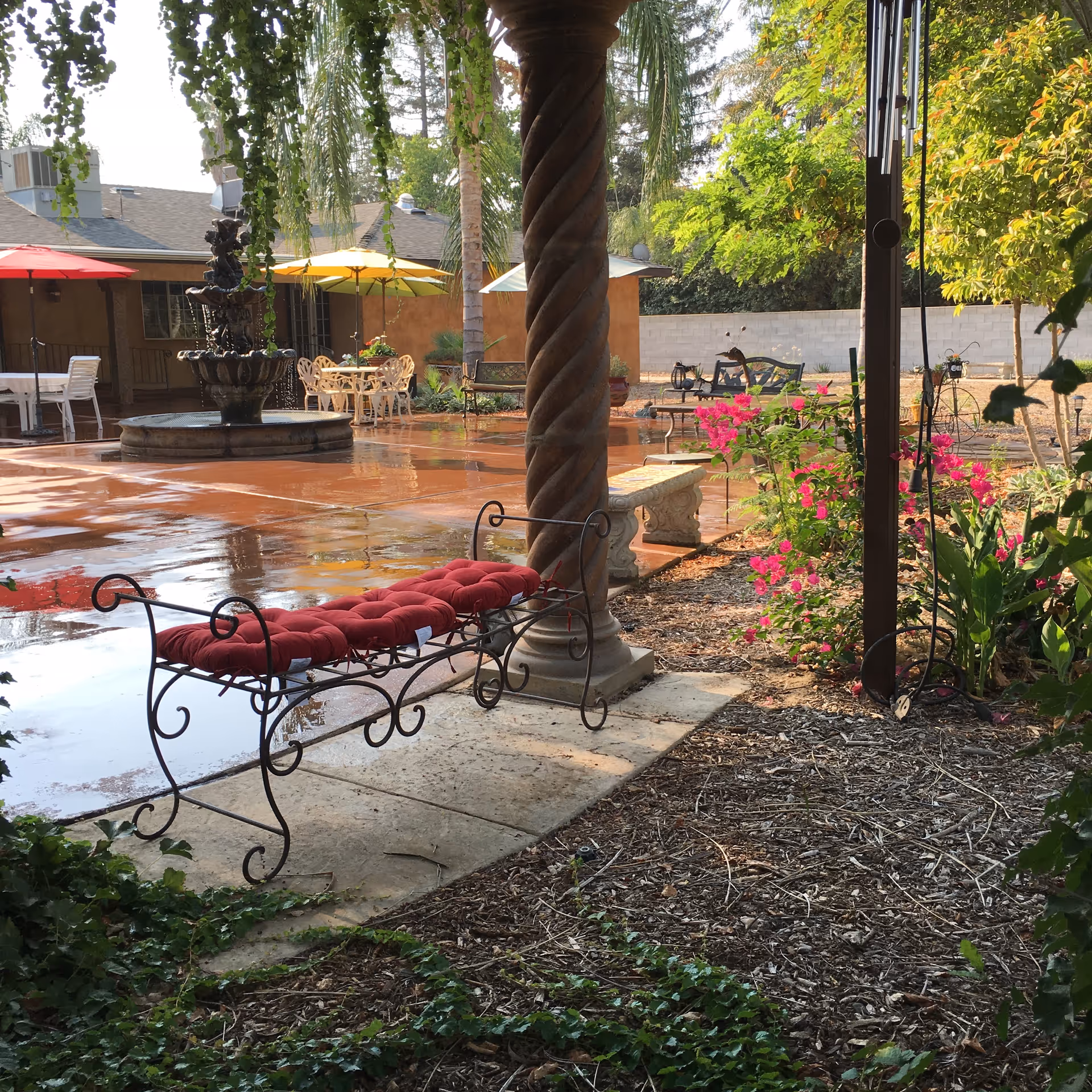 Outdoor patio area with a red cushioned wrought iron bench in the foreground, a stone pillar, pink flowering plants, and a water fountain surrounded by tables with colorful umbrellas in the background.