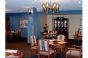 Dining area with several round tables and upholstered chairs, a chandelier overhead, and a china cabinet against blue-painted walls.