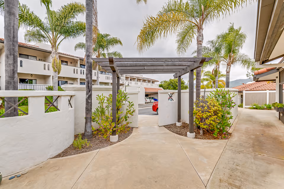 Outdoor courtyard area at Rancho Vista Senior Living featuring a paved walkway, a wooden pergola, palm trees, and white stucco walls with greenery and bushes on either side. Residential buildings with balconies and red-tiled roofs are visible in the background under a cloudy sky.