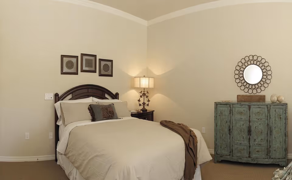A cozy bedroom with a neatly made bed featuring beige bedding and decorative pillows. There is a dark wooden headboard, a nightstand with a lamp, and three framed artworks above the bed. On the right side, there is a distressed teal cabinet with a round decorative mirror hanging above it. The walls are painted light beige and the room has a carpeted floor.
