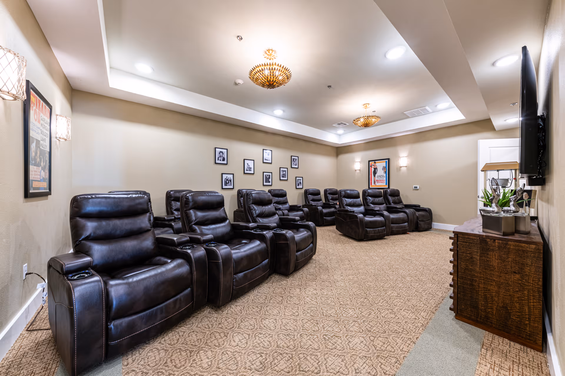 A cozy home theater room with two rows of black leather recliner chairs facing a wall-mounted flat screen TV. The room has beige walls decorated with framed pictures and movie posters, warm ceiling lights, and a wooden cabinet beneath the TV holding decorative items.