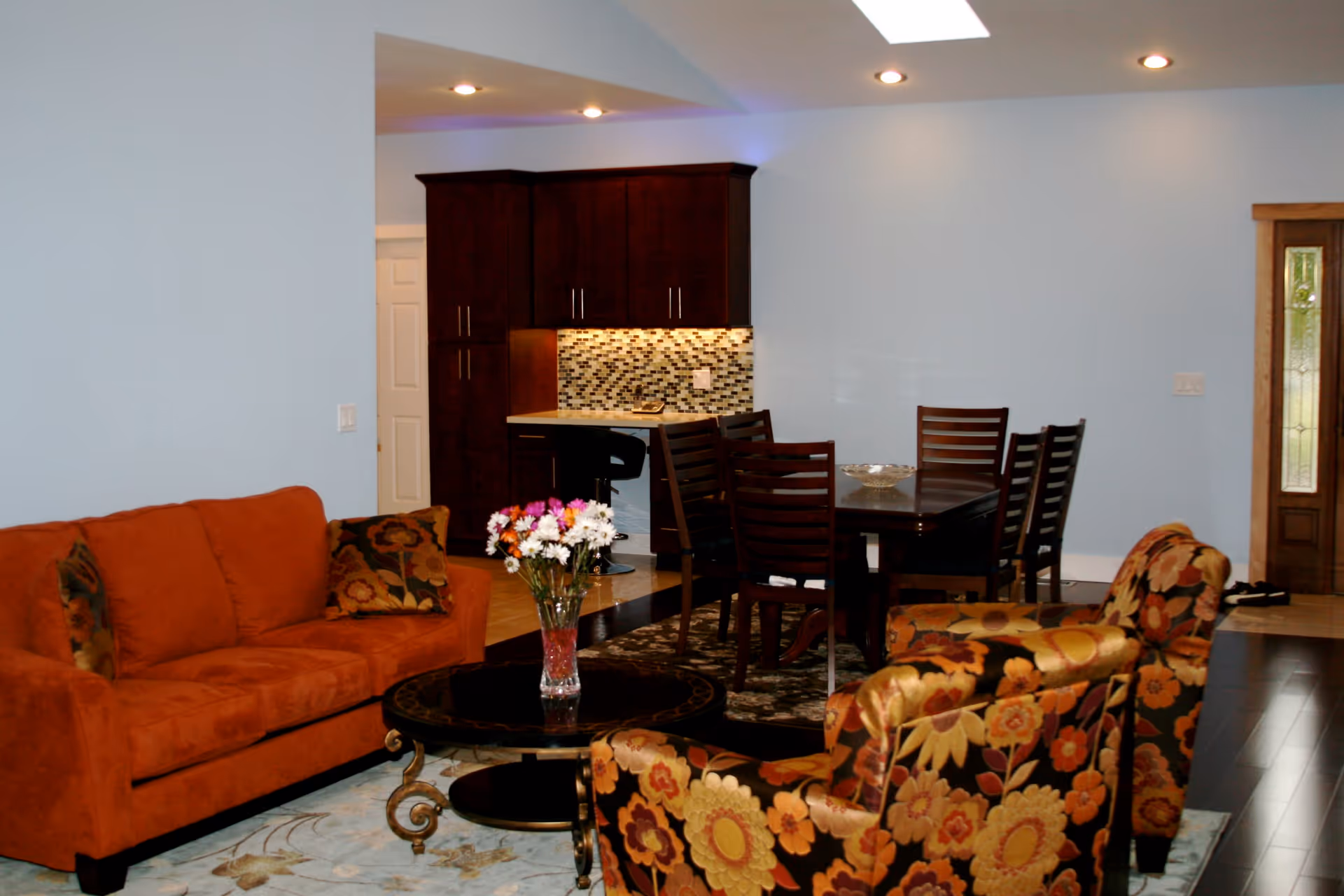 Open-plan living and dining area with an orange sofa, floral armchairs around a coffee table, and a dining table and kitchen in the background.