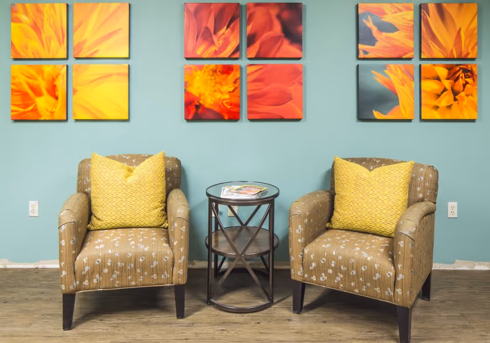 Two patterned armchairs with yellow cushions are placed on either side of a small round table with magazines on top. Behind the chairs, a teal wall features three sets of four square floral art pieces in shades of yellow, orange, and red. The floor is wooden.