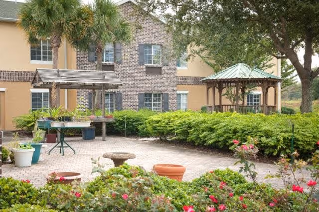Outdoor courtyard area of a senior living facility with a brick-paved patio, green shrubs, flowering plants, a wooden gazebo with a green roof, and a building with beige and brick exterior walls in the background.