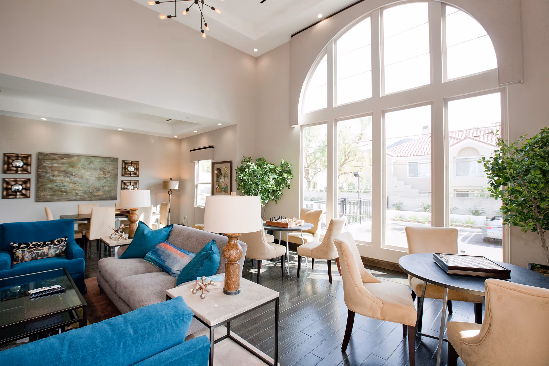 A bright and spacious living room area with large arched windows letting in natural light. The room features a gray sofa with blue pillows, blue armchairs, beige dining chairs around small round tables, and a chessboard on one table. There are decorative lamps, plants, and artwork on the walls, with dark wooden flooring and a high ceiling with recessed lighting.