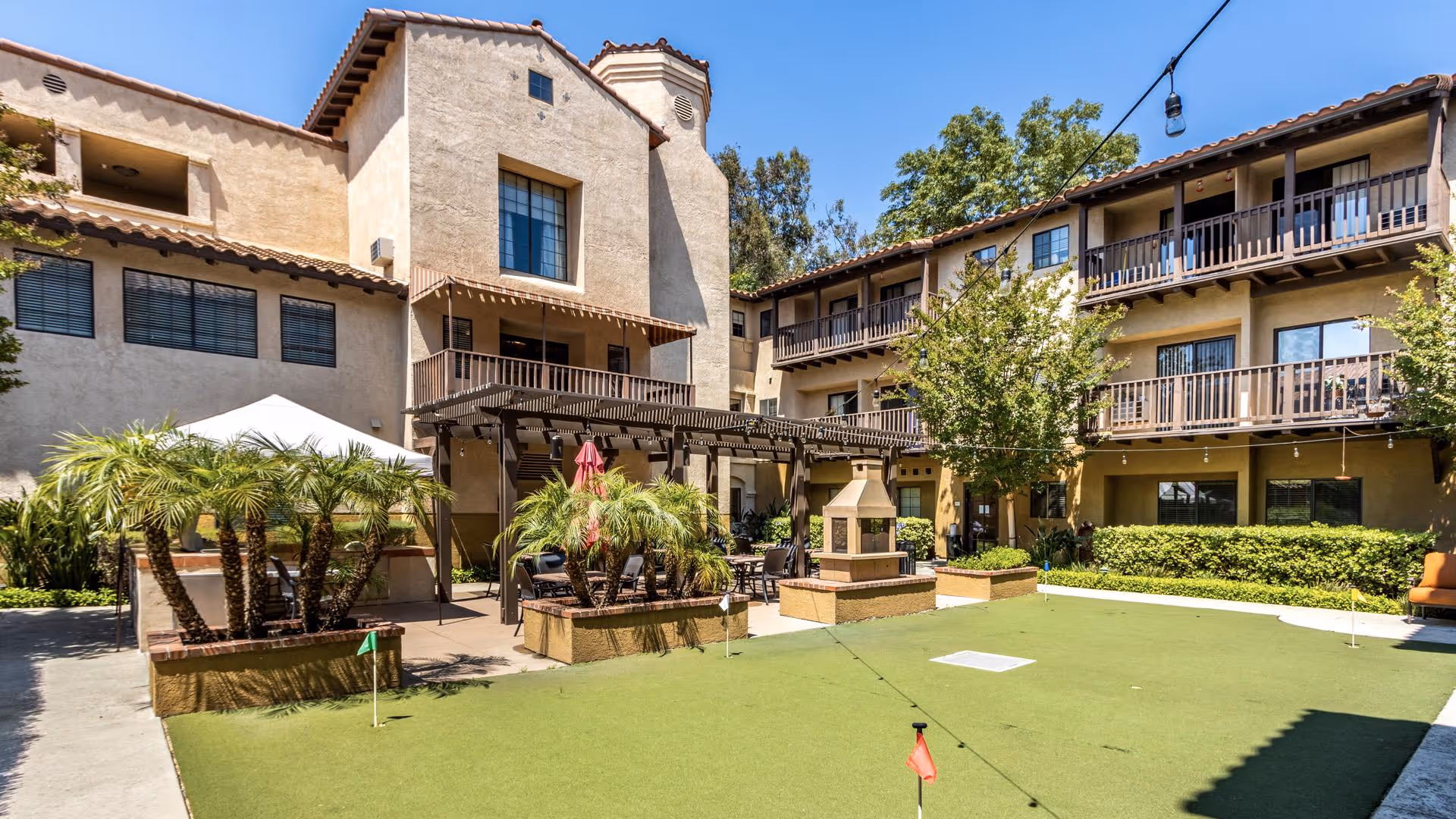 Outdoor courtyard area of a senior living facility with a green putting lawn, palm trees, a pergola with seating, and a multi-story building with balconies surrounding the space under a clear blue sky.