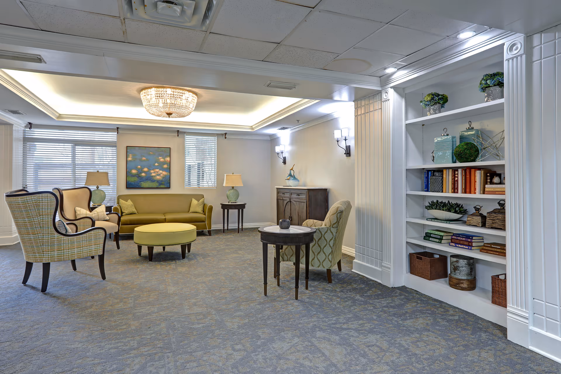 A well-lit living room area with a green sofa, two armchairs, a round ottoman, and side tables with lamps. There is a built-in white bookshelf filled with books and decorative items on the right side. The ceiling has recessed lighting and a chandelier, and the walls are painted light with two windows and a painting between them.