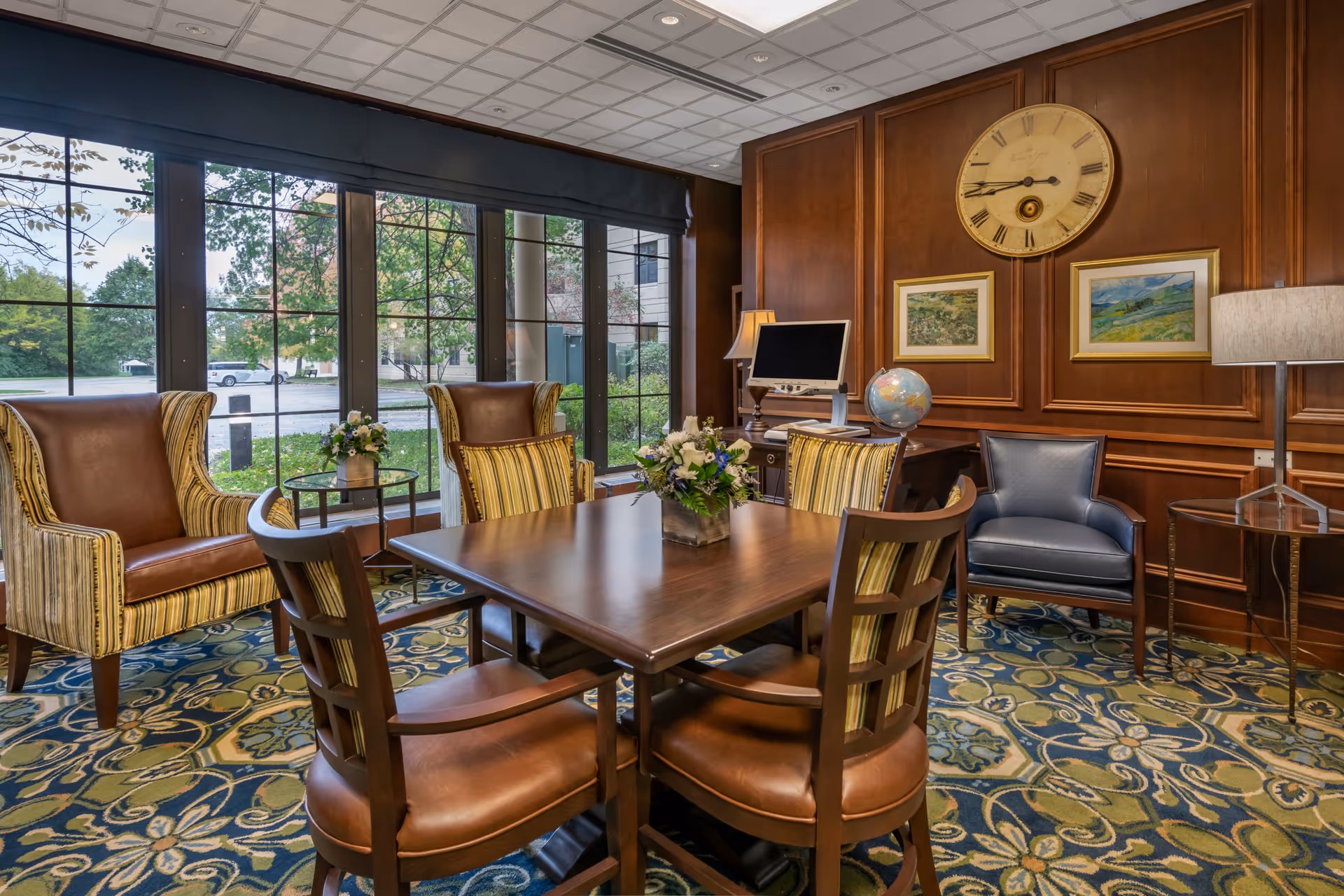 A cozy interior room with a wooden table surrounded by four chairs, two striped armchairs, and a blue armchair. The room features a large window with a view of greenery outside, a patterned carpet, wood-paneled walls with two framed paintings, a large round clock, a globe on a desk, and two lamps providing warm lighting.
