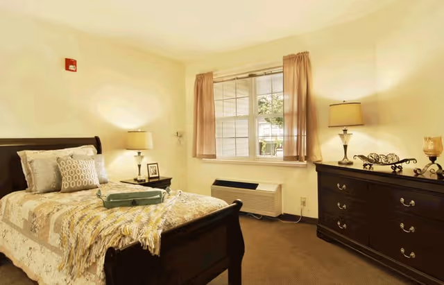 A cozy bedroom with a wooden bed frame, a bed covered with a patterned quilt and several pillows. There is a nightstand with a lamp and a framed photo next to the bed. A window with light pink curtains lets in natural light. Across from the bed is a dark wooden dresser with two lamps and decorative items on top. The room has beige walls and carpeted floor.