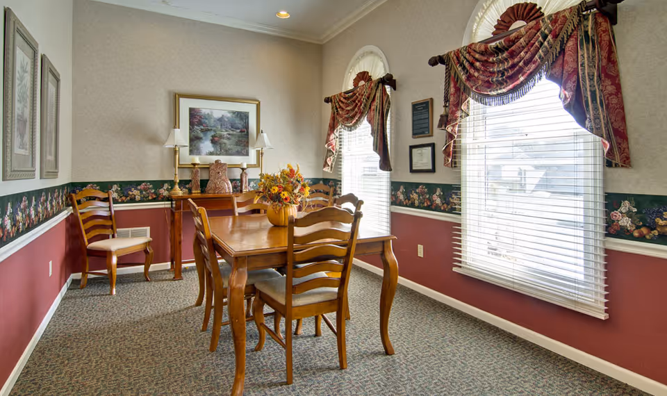 A dining room with a wooden table and six matching chairs. The walls are decorated with floral wallpaper border and two windows with red and gold valances. There are framed pictures and two lamps on a side table against the wall, and a floral centerpiece on the dining table.