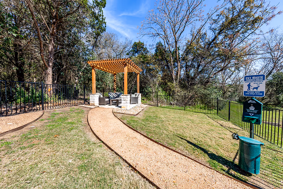 Outdoor area with a curved gravel pathway leading to a wooden pergola with cushioned chairs underneath. The area is surrounded by grass, trees, and a black metal fence. A pet waste station with a sign and trash bin is visible on the right side.