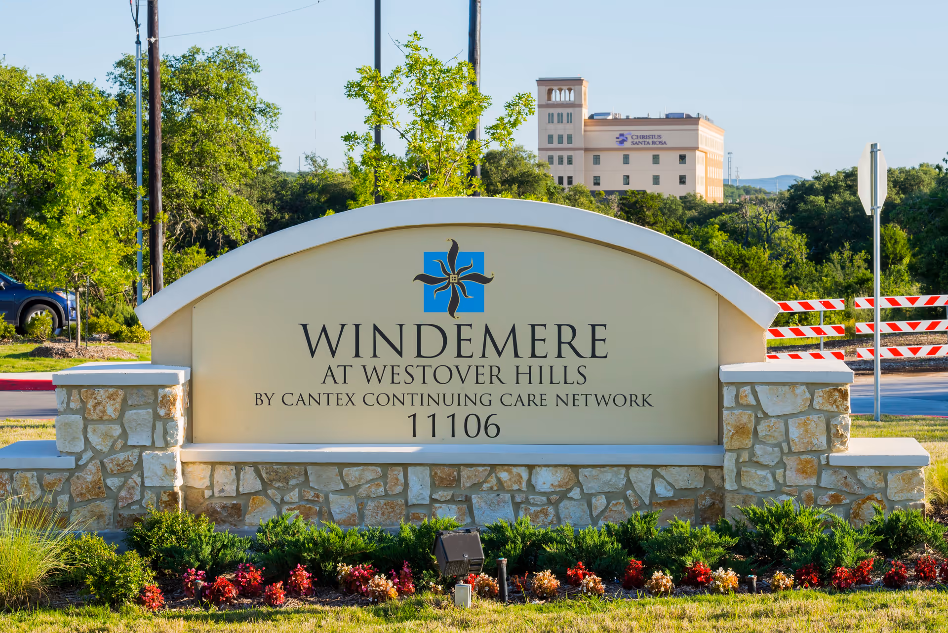 Stone and stucco sign for Windemere At Westover Hills by Cantex Continuing Care Network with the address 11106, surrounded by greenery and flowers, with a building and trees in the background.