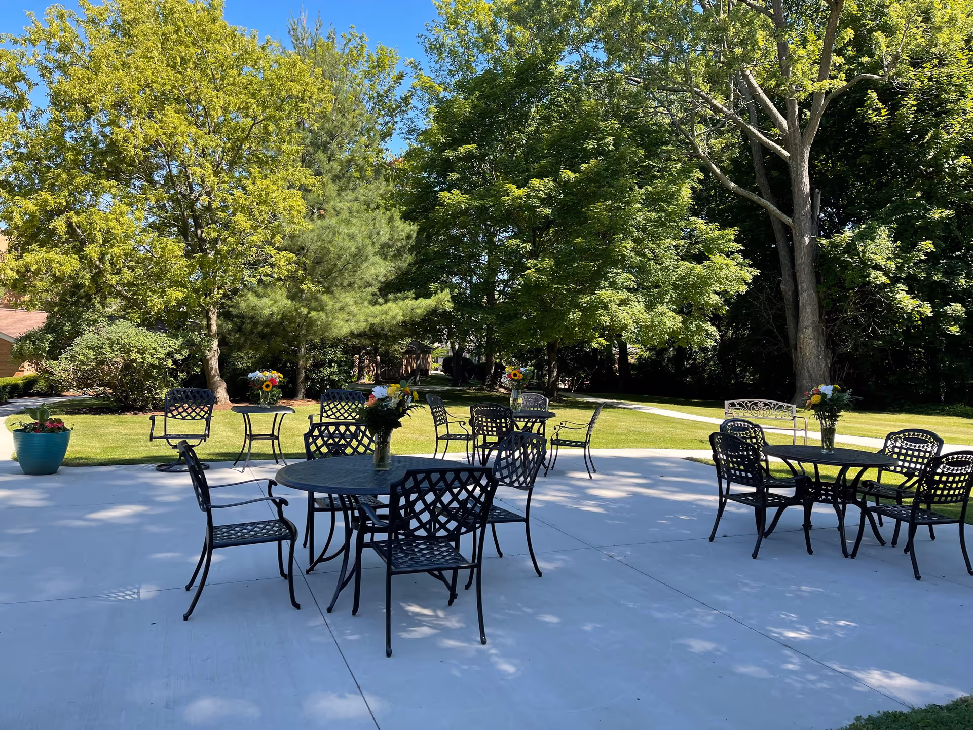 Outdoor patio area with several black metal tables and chairs arranged on a concrete surface. Each table has a vase with colorful flowers. The patio is surrounded by green grass, trees, and shrubs under a clear blue sky.