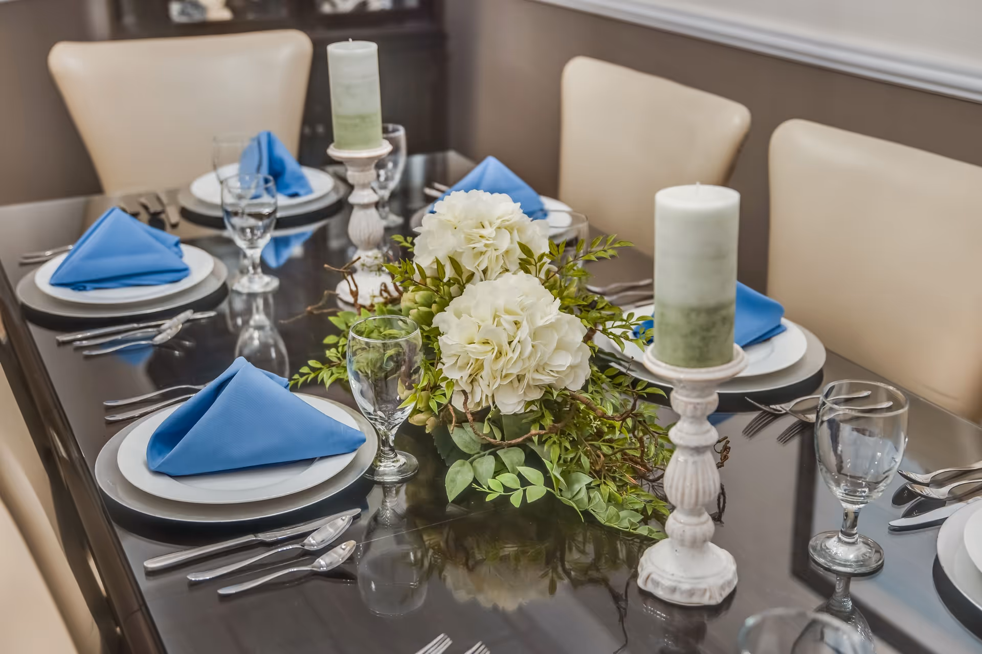 A formal dining table set with white plates, blue folded napkins, glassware, candles, and a white floral centerpiece.
