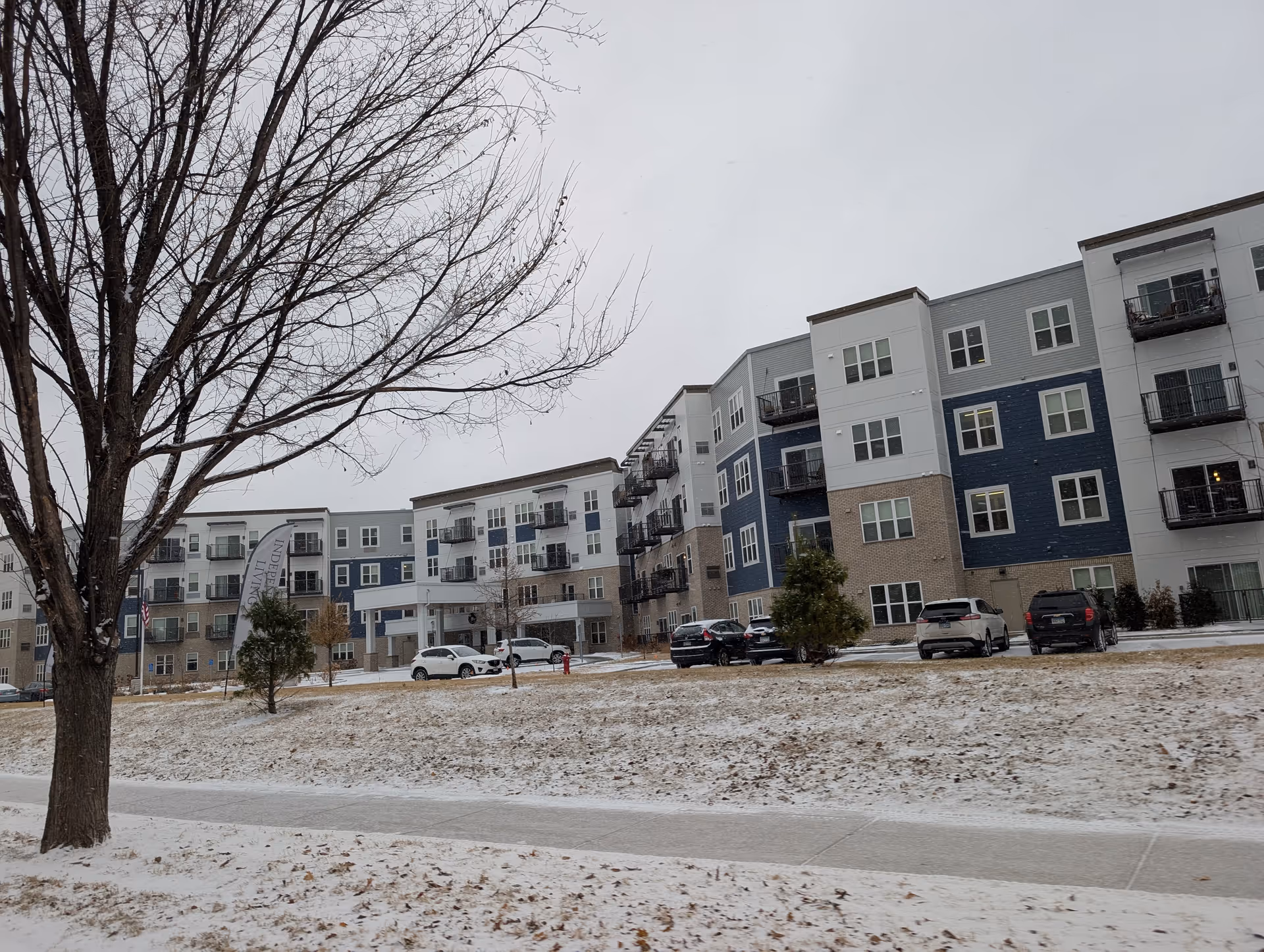 Exterior front view of a multi-story senior living apartment building with balconies, parked cars, a bare tree, and light snow on the ground.