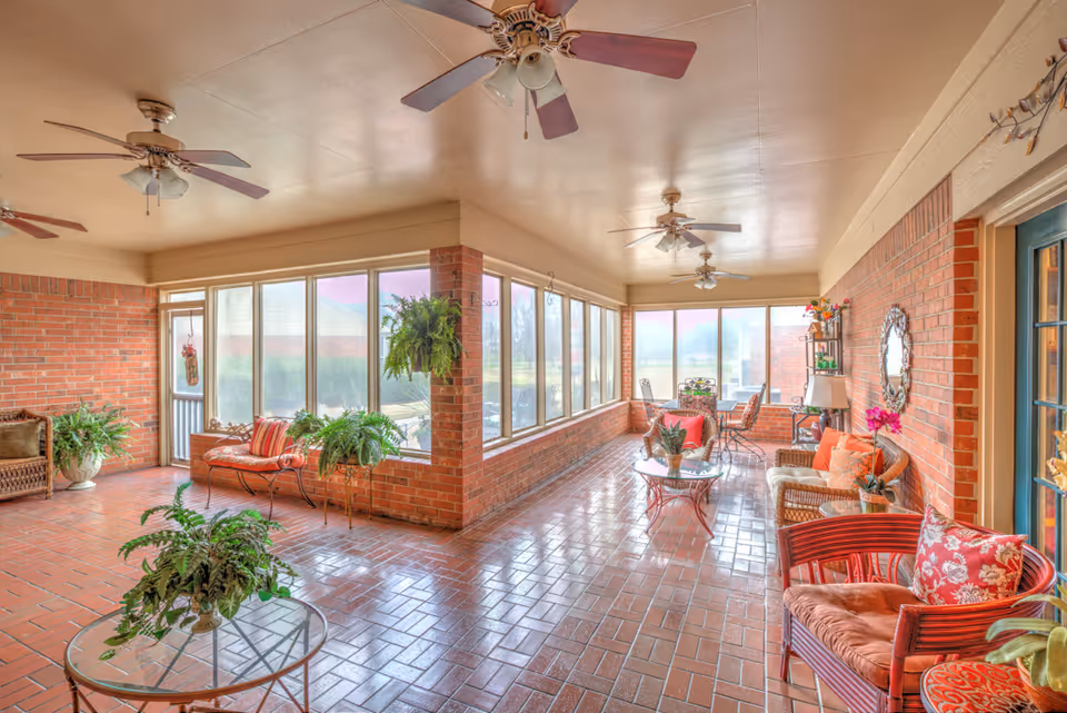 A spacious sunroom with brick walls and tiled floor featuring multiple ceiling fans. The room is furnished with various wicker and metal chairs, cushioned seating, glass-top tables, and decorated with several green plants and flowers. Large windows line the walls, allowing natural light to fill the space.