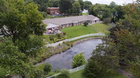 Aerial view of Stratford Rehabilitation Center showing a long single-story brick building, a curved pond, white fencing, lawns, and surrounding trees.