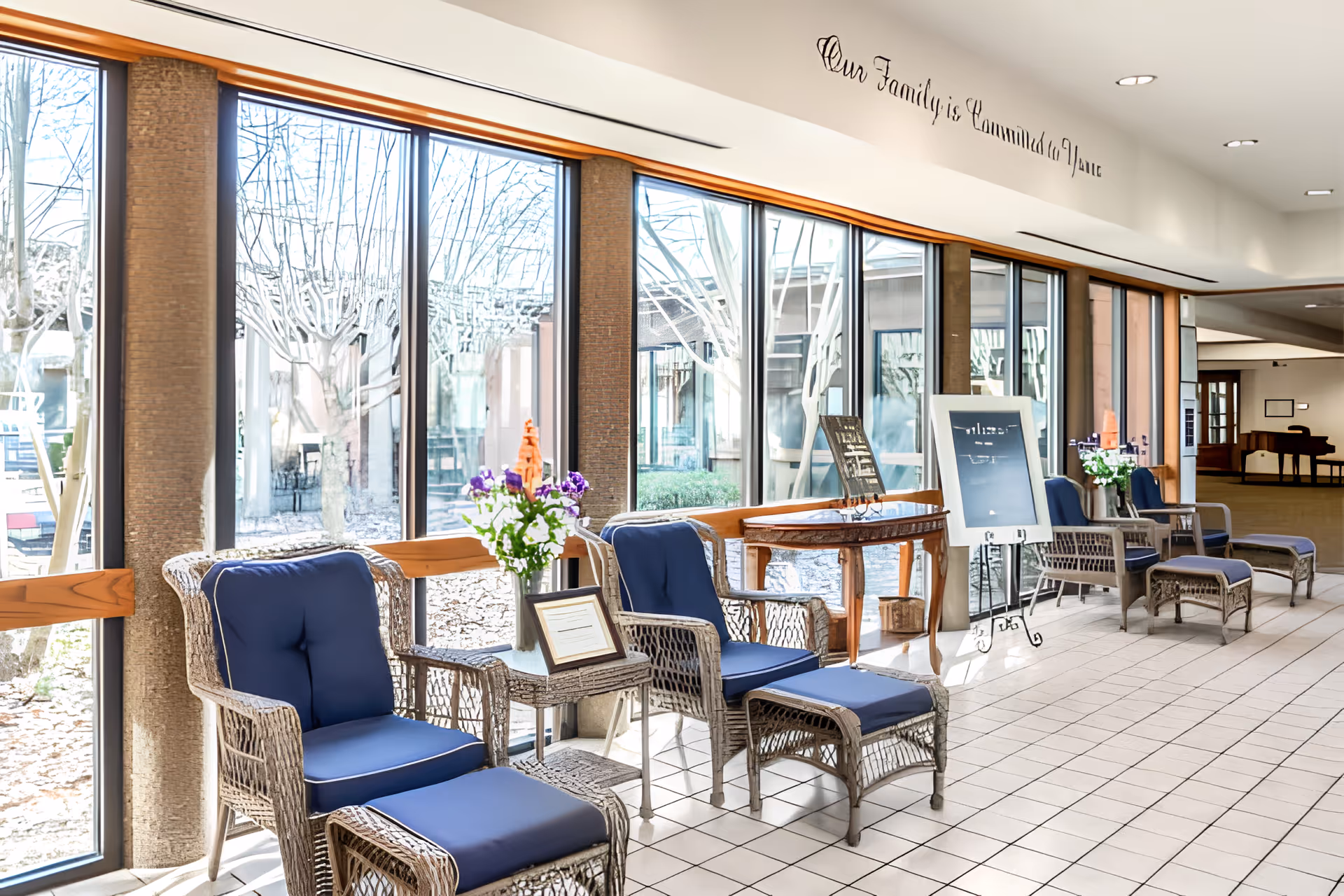 A bright and spacious indoor seating area with large floor-to-ceiling windows showing a view of leafless trees outside. The seating consists of wicker chairs with blue cushions and matching ottomans arranged along the windows. Small tables with flower arrangements and framed signs are placed between the chairs. The floor is tiled in white, and a wooden table with decorative items is visible further down the corridor. On the wall above the windows, there is a decorative script that reads 'Our Family is Committed to You'.