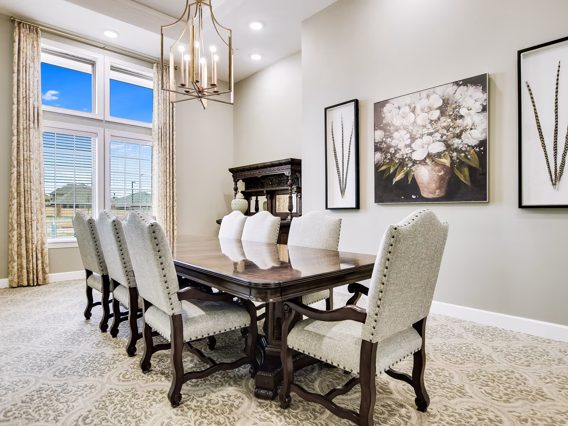 A bright dining room with a large wooden dining table surrounded by eight upholstered chairs. The room features a large window with beige curtains, a decorative chandelier, and three framed artworks on the wall, including a painting of white flowers in a vase.