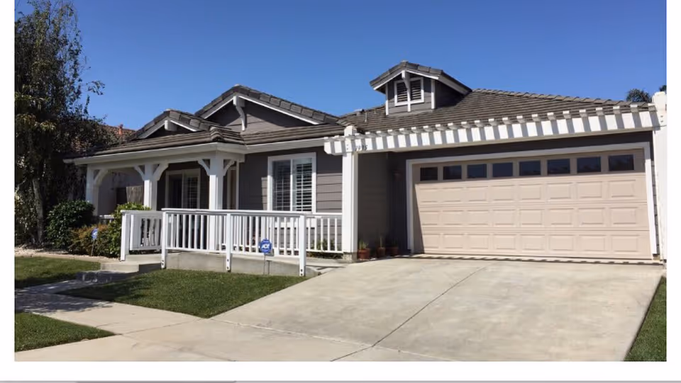 Exterior view of a single-story residential building with a gray facade, white trim, a covered porch with white railings, and a two-car garage. The driveway and a small lawn are visible under a clear blue sky.