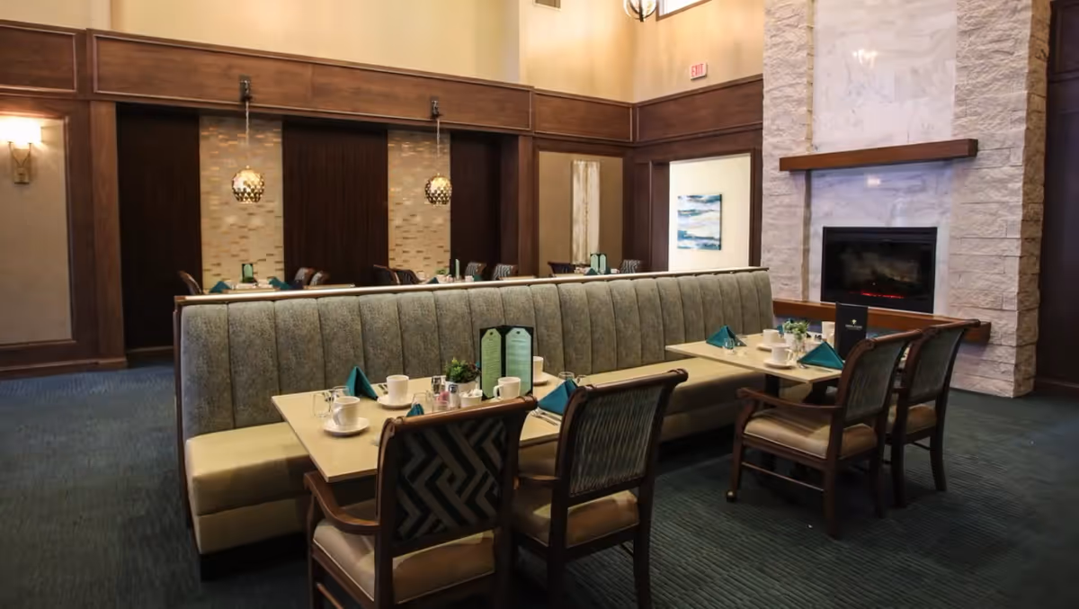 A dining area with a long upholstered bench and wooden chairs around tables set with cups, glasses, napkins, and menus. The room features a stone fireplace, wood paneling, pendant lights, and carpeted floor.