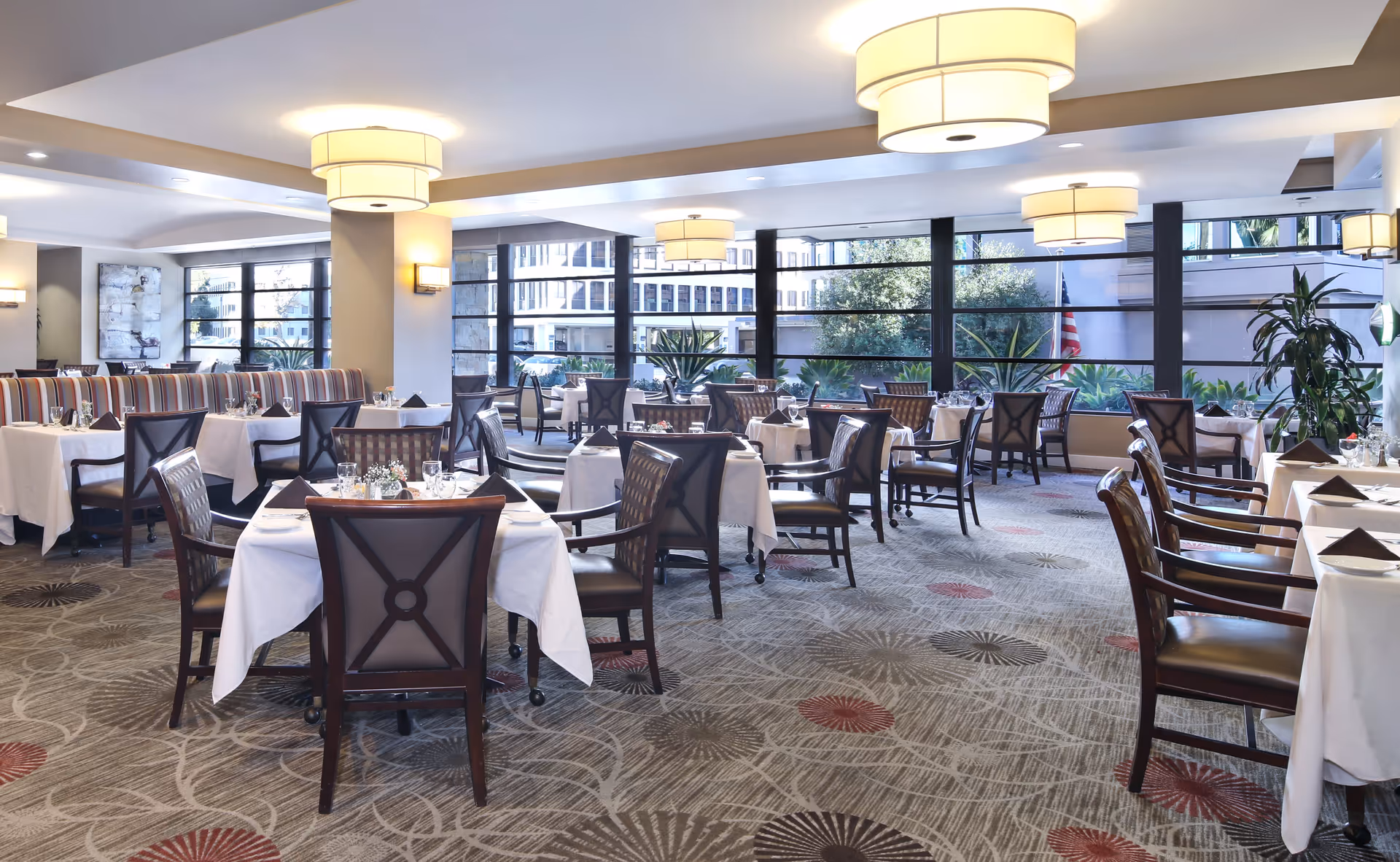 Bright and spacious dining room with multiple tables covered in white tablecloths, set with glassware, napkins, and silverware. The room features large windows letting in natural light, modern ceiling lights, patterned carpet, and a mix of wooden chairs and booth seating along one wall.