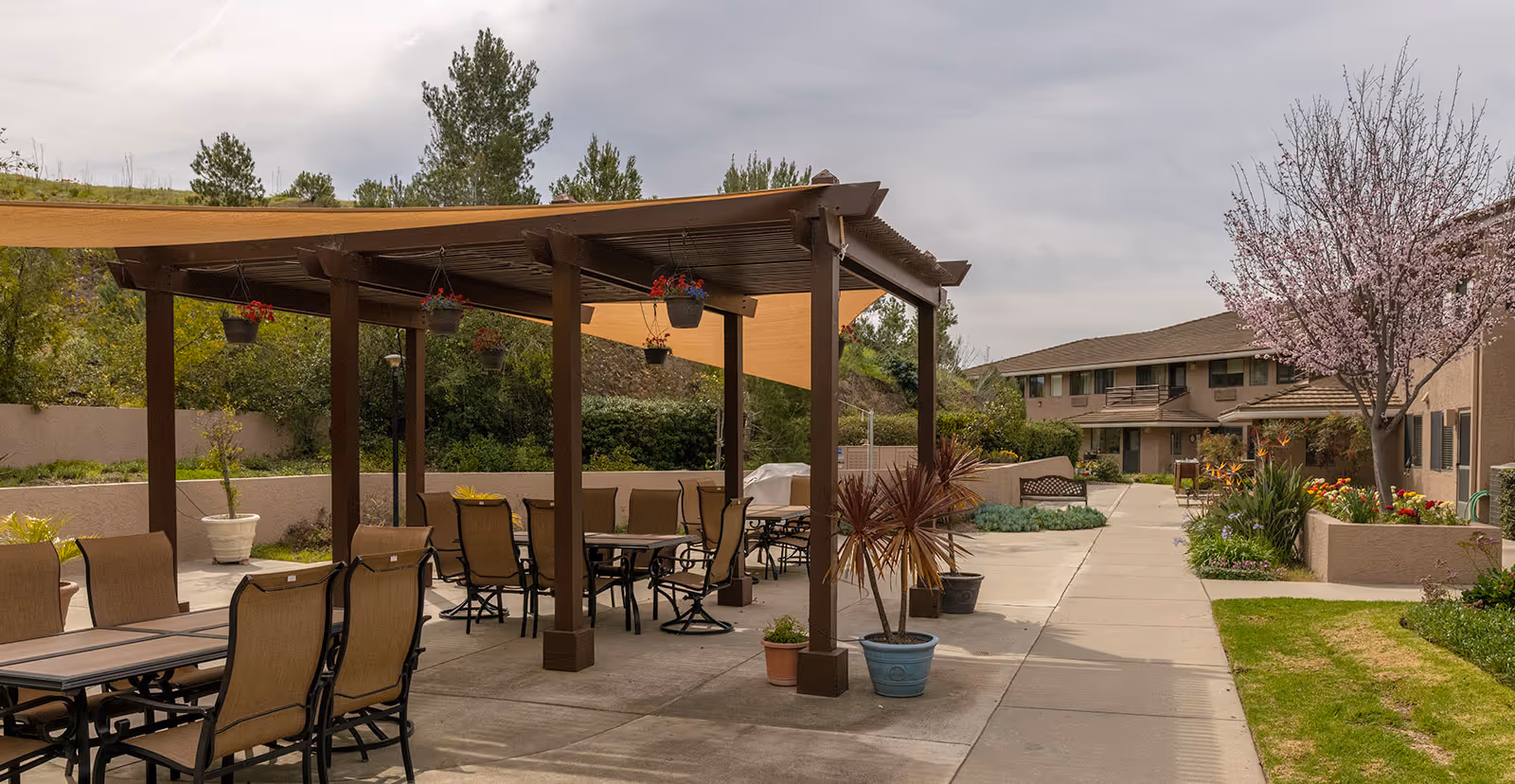 Outdoor patio area at Las Brisas Retirement Community with tables and chairs under a wooden pergola, hanging flower pots, potted plants, a walkway, and a blooming tree near residential buildings.