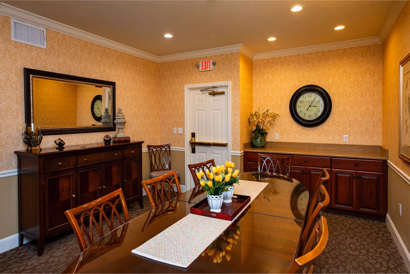A dining room with a polished wooden table surrounded by wooden chairs. The table has a runner and a tray with three white pots of yellow tulips. The walls are decorated with patterned wallpaper in warm tones. There is a large mirror above a wooden sideboard on the left wall, and a round clock on the right wall above a countertop with cabinets. A white door with an exit sign above it is visible at the back of the room.