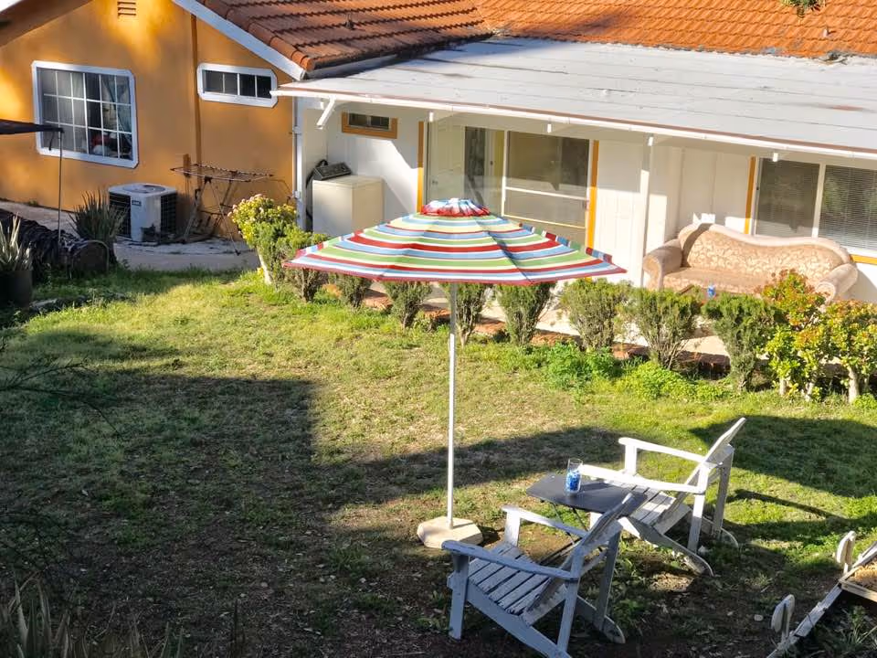 Outdoor garden area with green grass, two white wooden chairs, a small table with a blue glass on it, and a colorful striped umbrella providing shade. In the background, there is a yellow building with a tiled roof, windows, a beige couch on a porch, and some bushes along the edge of the porch.