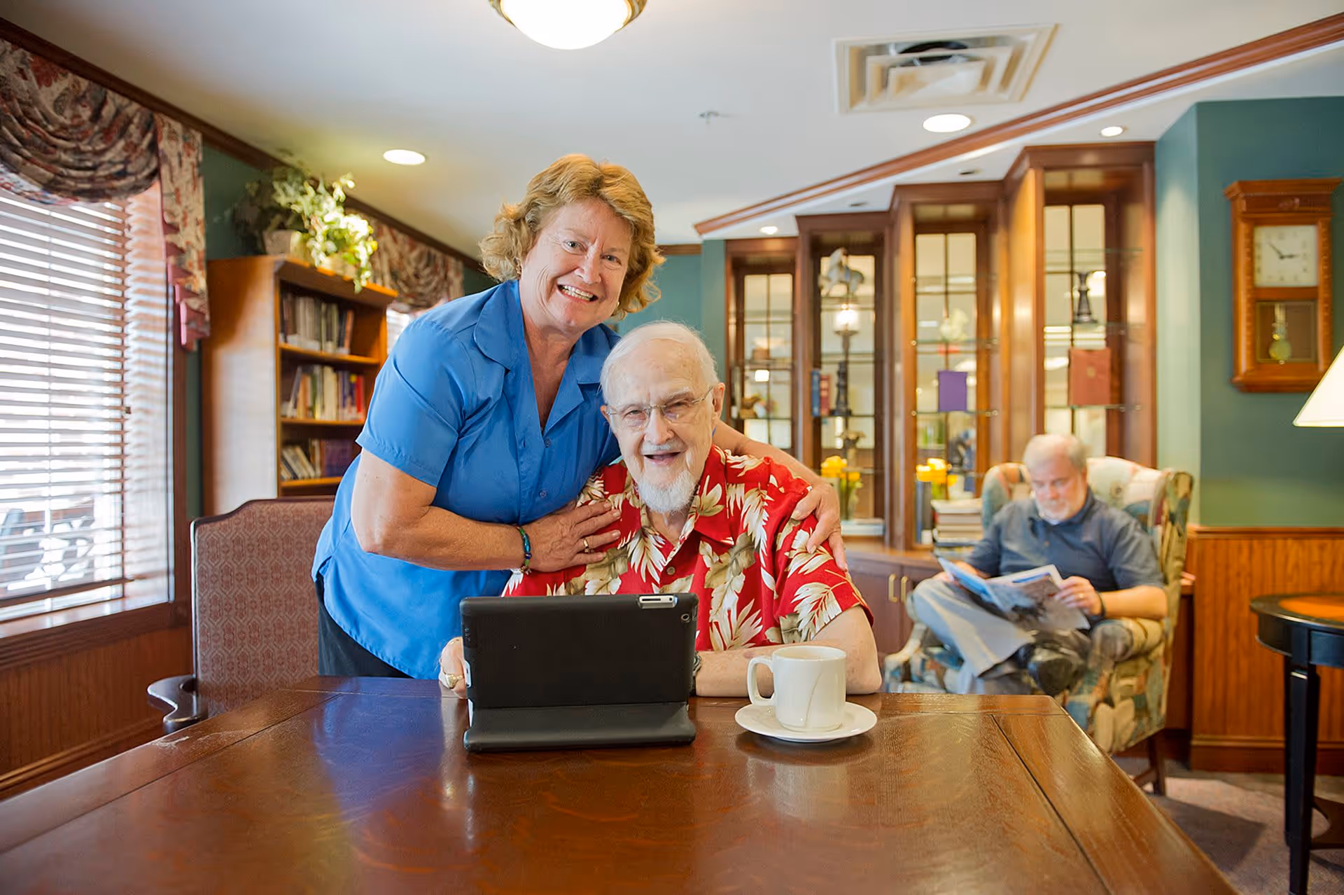 An elderly man wearing a red floral shirt sits at a wooden table with a tablet and a cup of coffee in front of him. A woman in a blue shirt stands behind him, smiling and resting her hands on his shoulders. In the background, another elderly man is seated in a patterned armchair reading a magazine. The room has wooden furniture, a bookshelf, a glass display cabinet, and a wall clock.