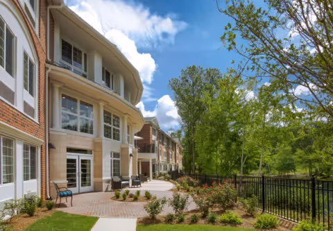 Exterior view of a multi-story brick senior living building with a paved patio, seating, landscaped grounds, and trees beside a fenced pond.