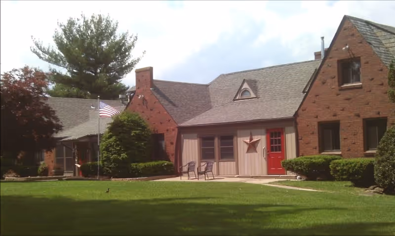 Front view of a brick and wood-faced building with a red door, American flag, patio chairs and a grassy lawn.