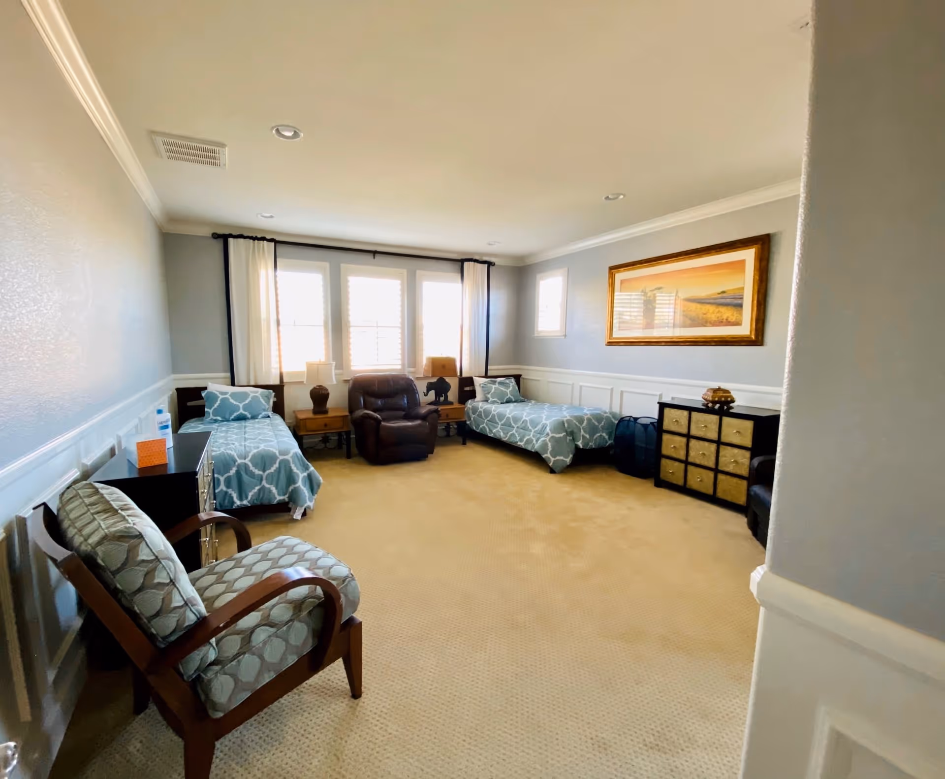 A bright and spacious bedroom with two twin beds covered in blue and white patterned bedding. Between the beds is a brown leather recliner chair and two wooden nightstands with lamps. There is a cushioned armchair with a wooden frame in the foreground. The walls are painted light gray with white wainscoting, and a large framed landscape painting hangs on the wall above a black dresser with multiple drawers. The room has beige carpet and three large windows with white curtains letting in natural light.