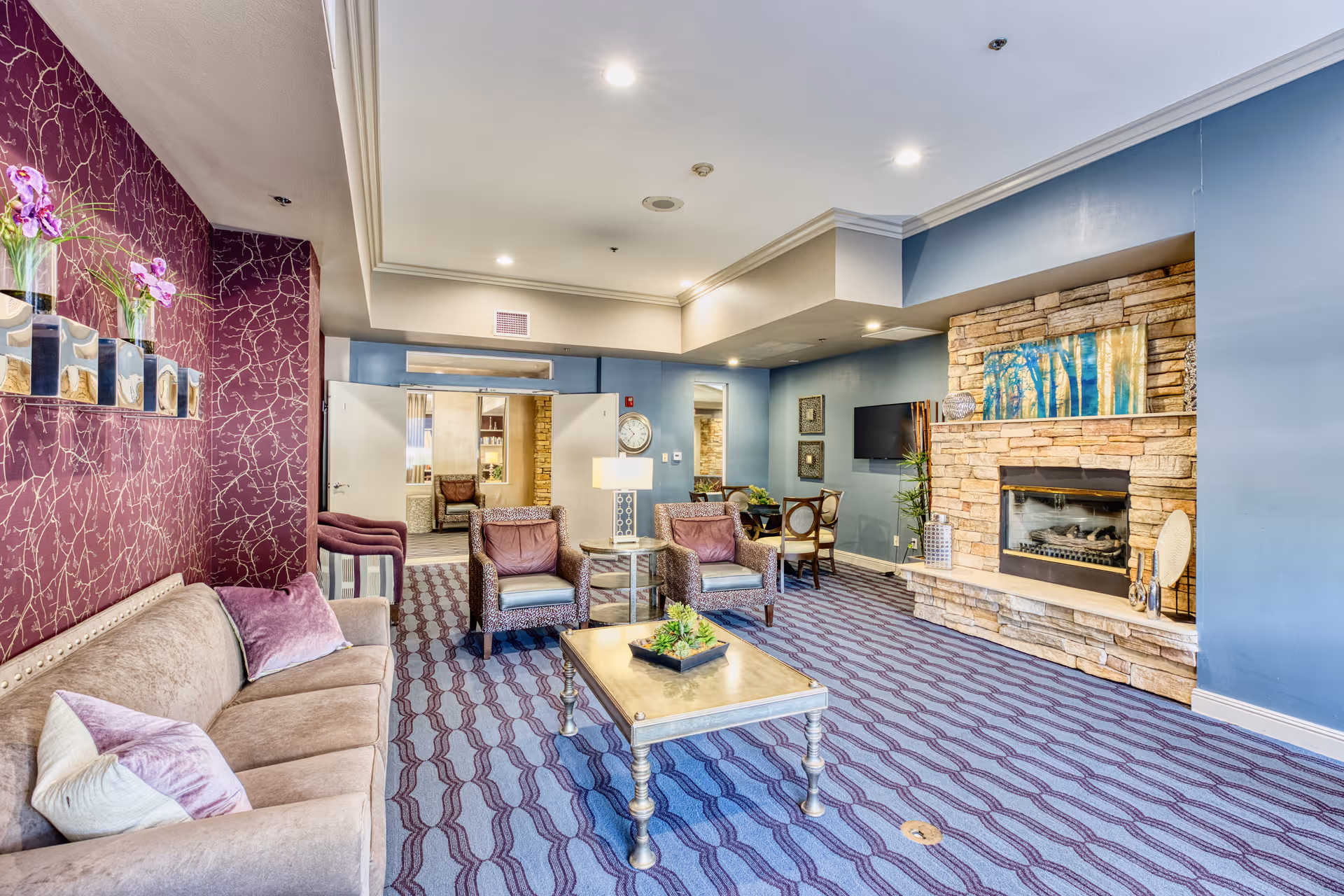 A spacious living room area with a beige sofa and purple pillows on the left, two armchairs with a small round table between them, a rectangular coffee table with a plant centerpiece in the middle, and a stone fireplace with a colorful painting above it on the right. The walls are painted blue and purple with a patterned carpet on the floor. There is a TV mounted on the wall and additional seating and decor in the background.