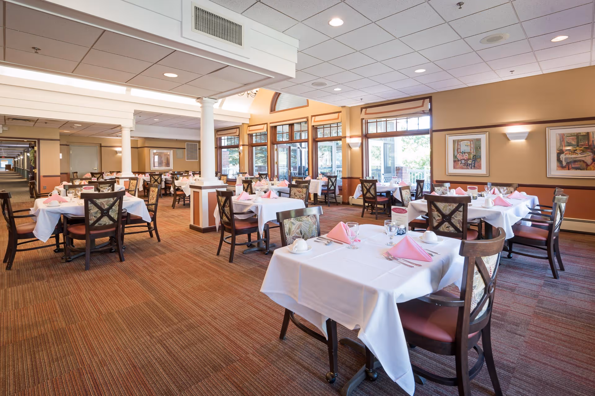 Spacious dining room with multiple tables set with white linens and pink napkins, wooden chairs, and large windows letting in natural light.