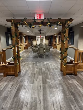 Indoor hallway with wood-look flooring and a wooden arch decorated with green vines and yellow flowers. There are chairs and tables arranged in the middle of the hallway, with wooden benches on either side. The ceiling has recessed lighting and an illuminated exit sign above the arch.