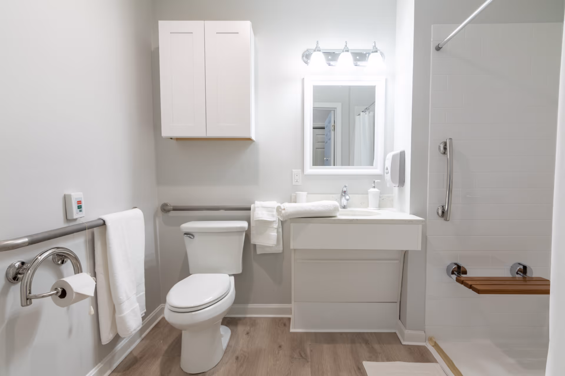 A clean, accessible bathroom with a white toilet, a white vanity with a sink, a mirror with three lights above it, a wall-mounted cabinet, grab bars near the toilet and in the shower, a towel rack with a white towel, and a shower area with a wooden fold-down seat and a white shower curtain.