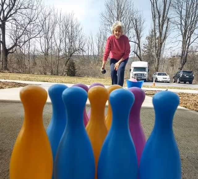 An elderly woman wearing a pink sweater and dark pants is playing an outdoor bowling game with colorful plastic pins arranged in front of her. The background shows leafless trees, a grassy area, and parked vehicles under a clear sky.
