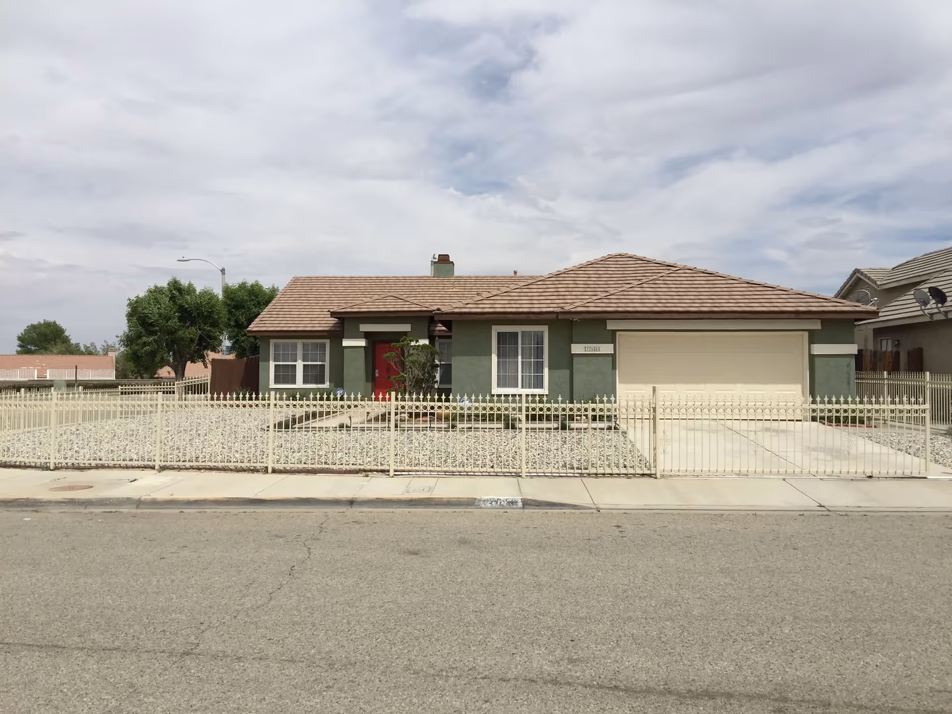 Single-story house with a beige tiled roof, green exterior walls, a red front door, and a two-car garage. The front yard is covered with gravel and enclosed by a cream-colored metal fence. There are a few small bushes near the entrance and a tree to the left side of the house. The sky is partly cloudy.