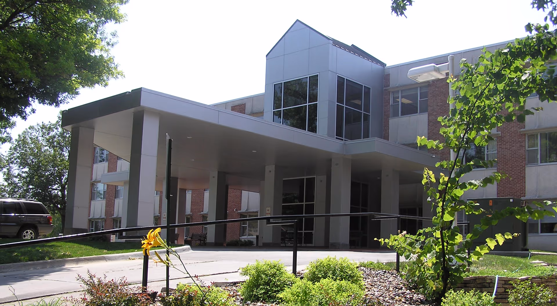 Front entrance of a multi-story brick senior living building with a covered porte-cochere and landscaped driveway.