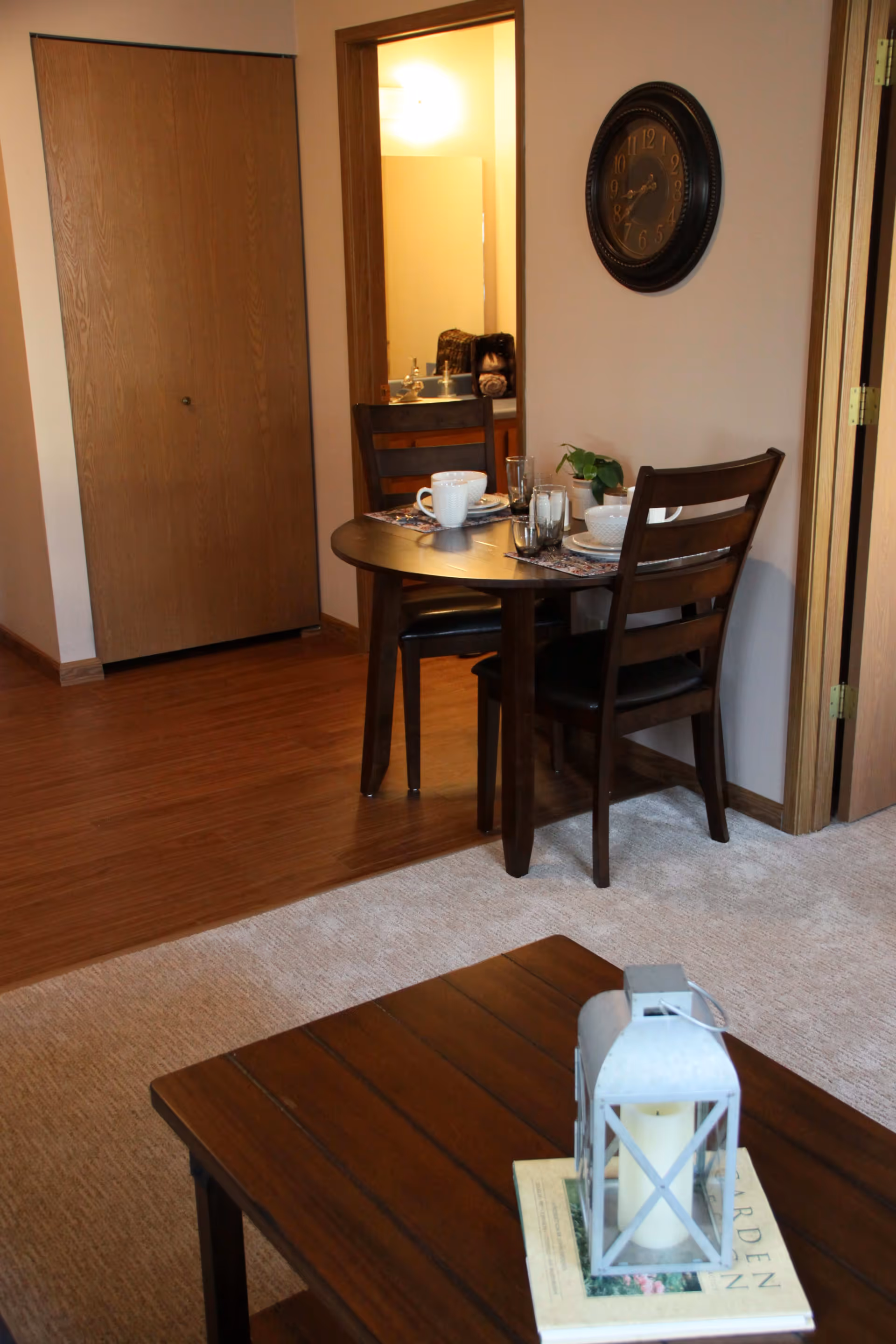 Small dining area with a round wooden table set for two, chairs, a wall clock, and a coffee table with a lantern in the foreground.