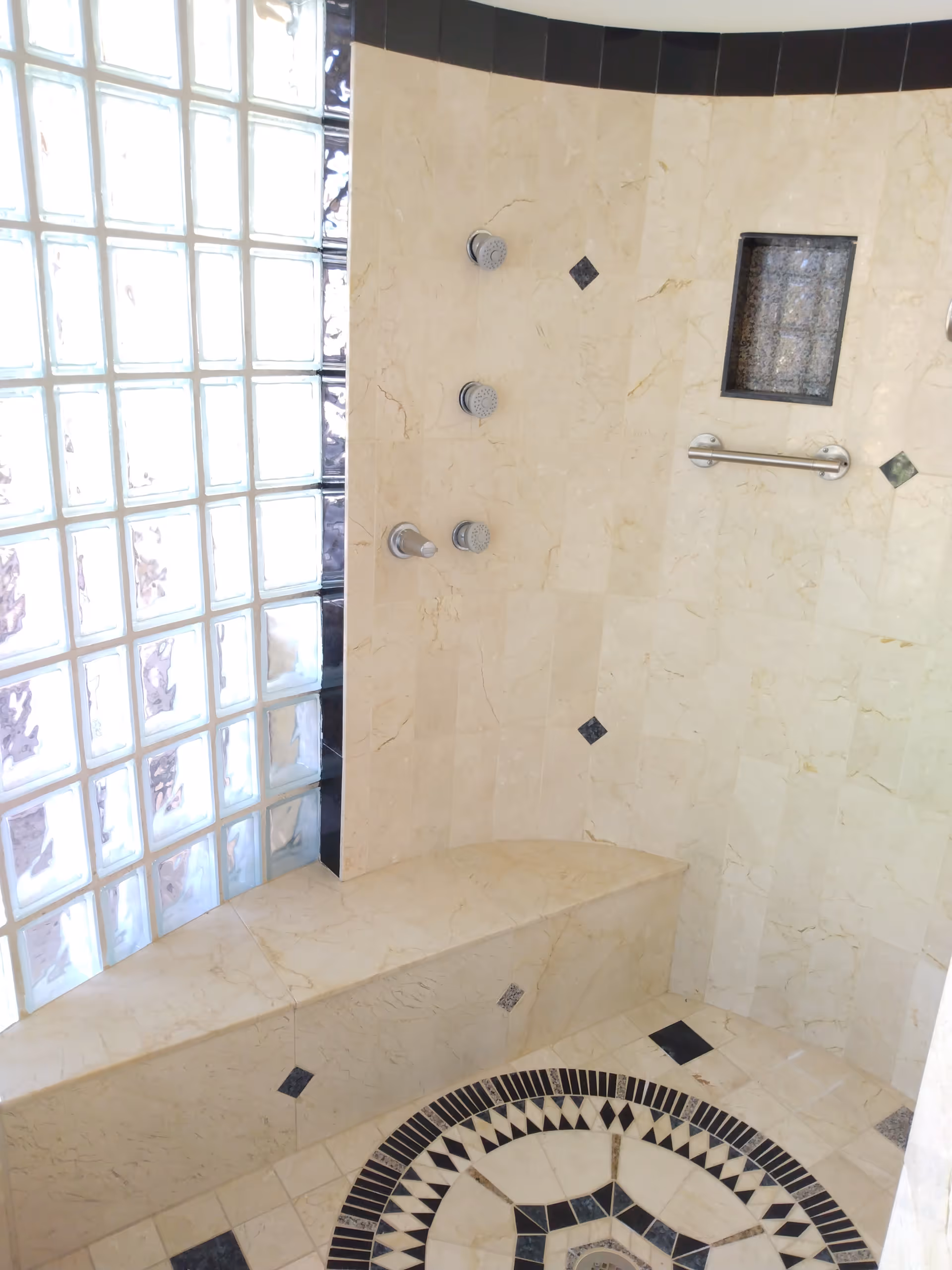A tiled shower area with beige marble walls and floor featuring black diamond accents. The shower has three shower heads mounted on the wall, a built-in bench, a small recessed shelf, and a glass block window allowing natural light to enter.