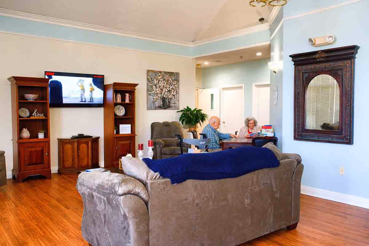 A cozy living room in Belmont Assisted Living with a gray couch covered by a blue blanket, wooden shelves with decorative items, a wall-mounted TV, a large mirror with an ornate wooden frame, and two elderly people sitting at a table in the background.