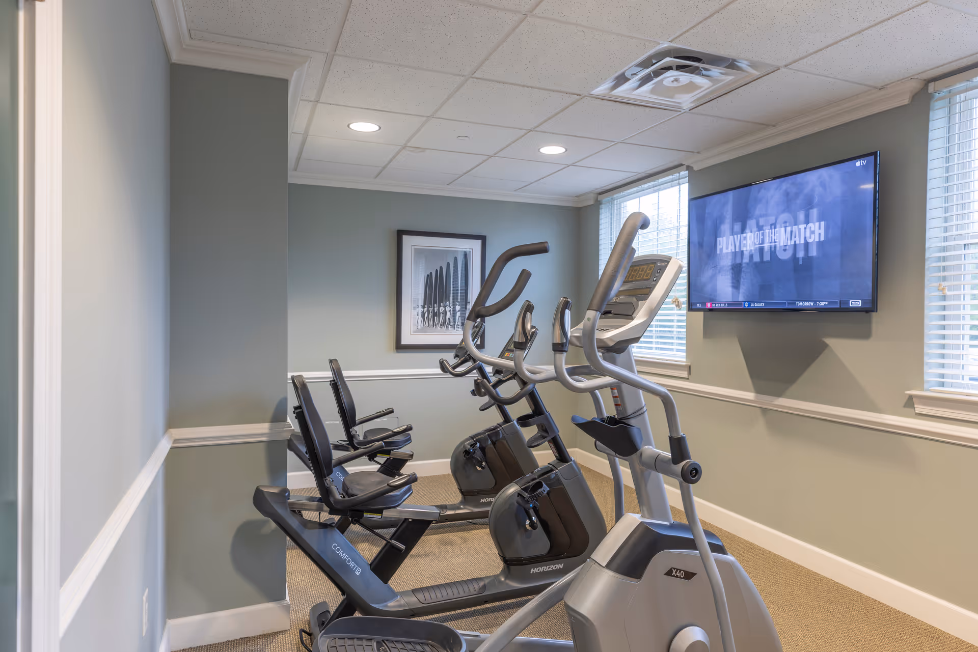 A small fitness room with exercise bikes and elliptical machines facing a wall-mounted TV displaying 'Player of the Match'. The room has light green walls, two windows with blinds, a framed picture on the wall, and recessed ceiling lights.