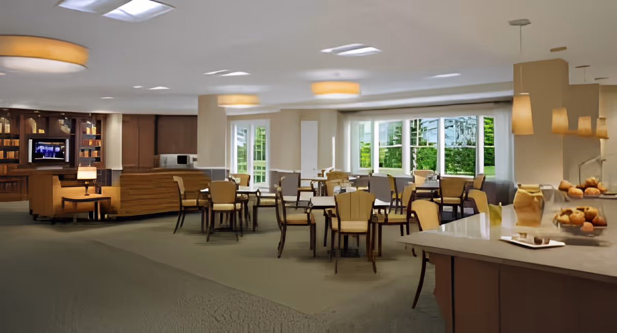 A spacious dining room in an assisted living facility with multiple tables and chairs arranged neatly. Large windows allow natural light to brighten the room, and there is a counter area with baked goods displayed. The room has warm lighting fixtures and a cozy atmosphere.