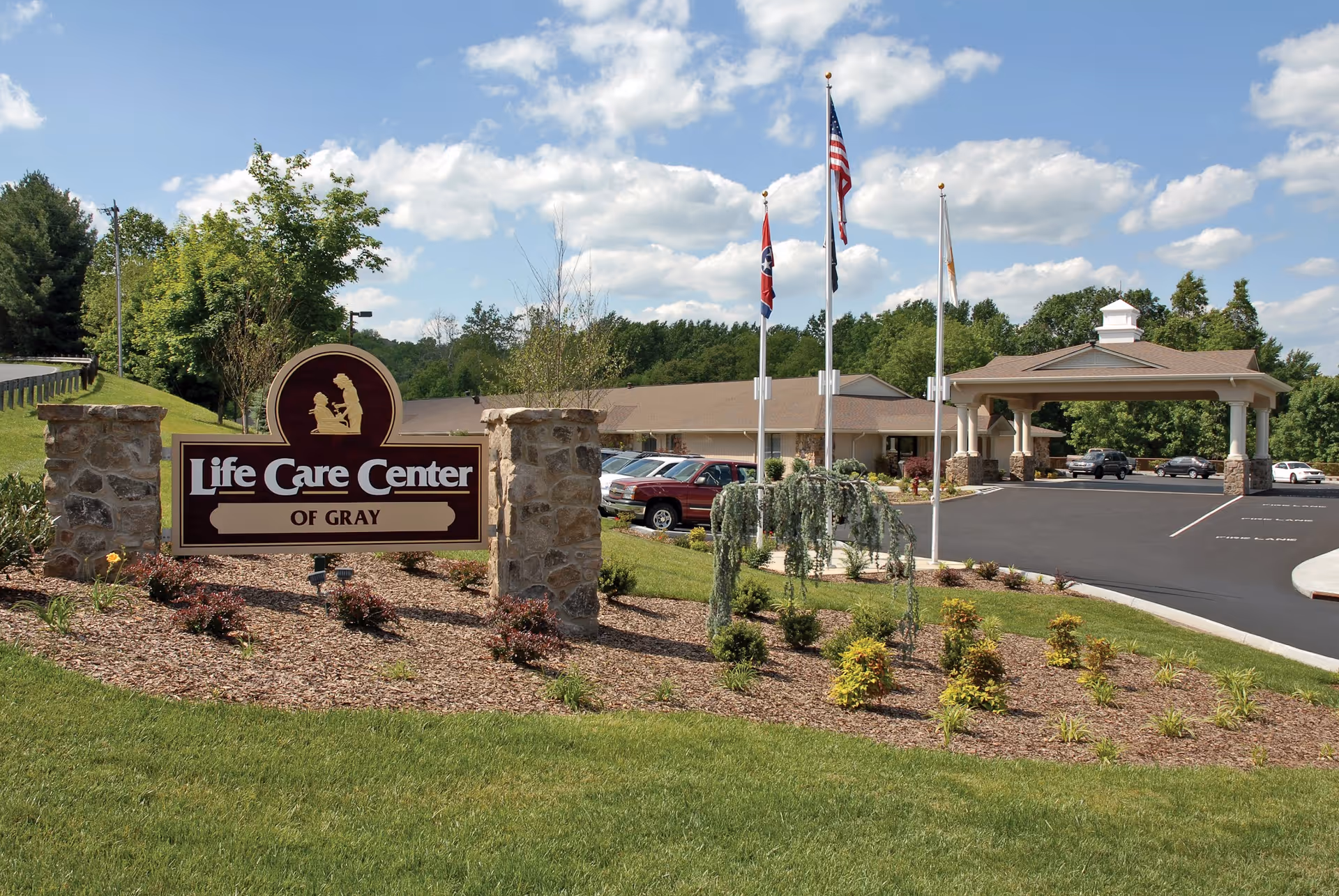 Exterior view of Life Care Center of Gray showing the facility sign mounted on stone pillars, a landscaped garden with small bushes and trees, a parking lot with several cars, a covered entrance, and three flagpoles with flags against a partly cloudy sky.