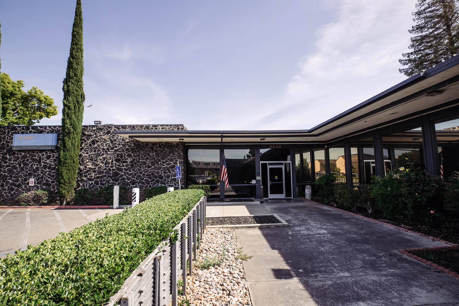 Front entrance of a low, single-story nursing facility with a stone facade, glass doors, an American flag, and a landscaped driveway.