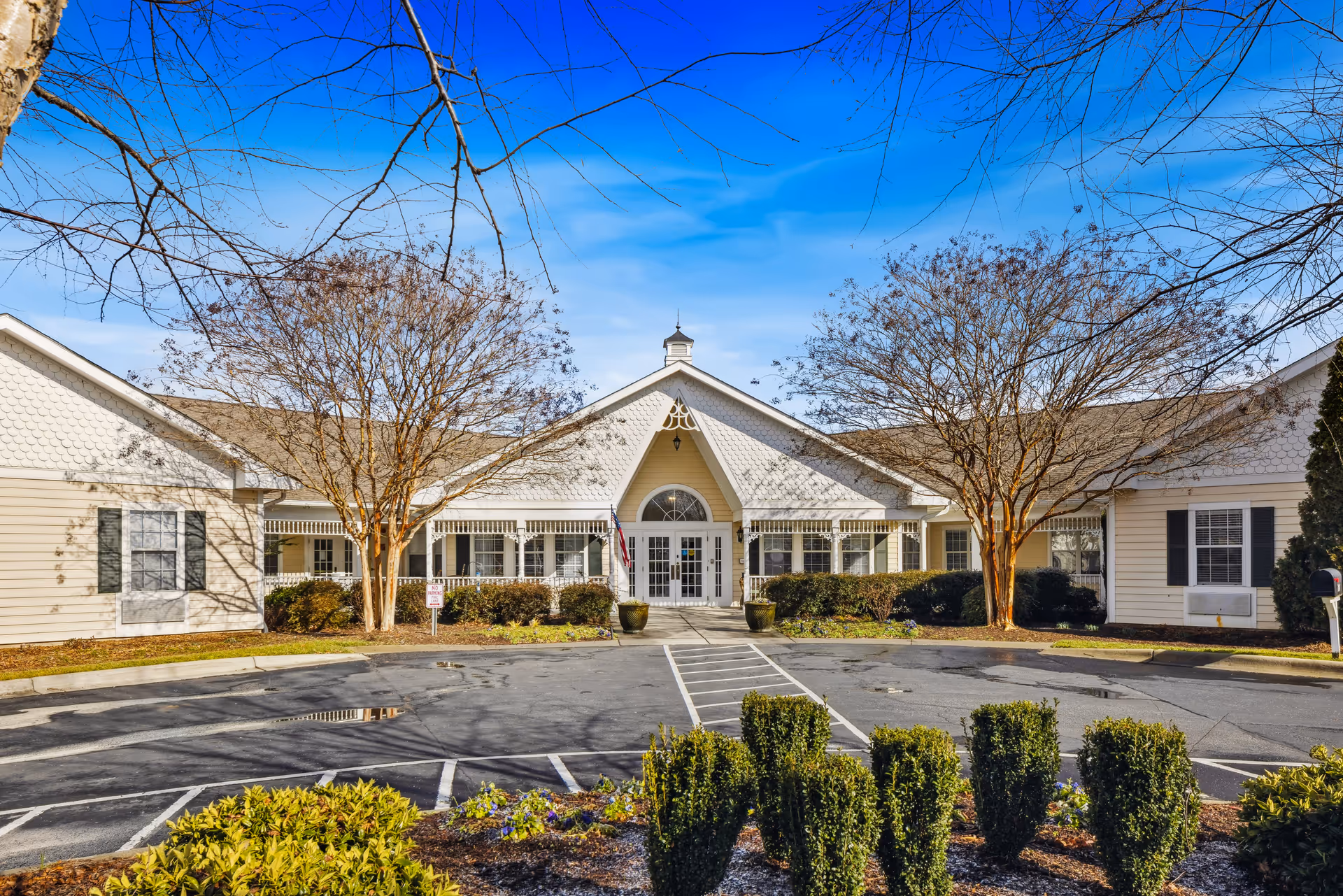 Front exterior view of TerraBella Indian Trail facility showing a single-story building with white siding and a peaked roof. There are two leafless trees near the entrance, shrubs along the building, a paved parking area with marked spaces, and a clear blue sky above.
