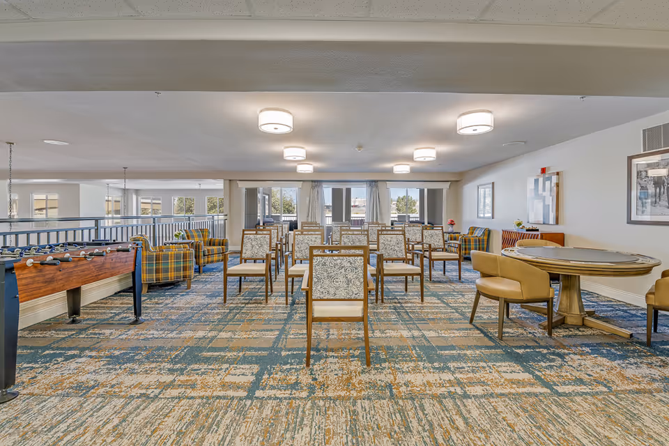 A spacious common area in a senior living facility featuring multiple rows of wooden chairs with patterned cushions arranged in the center. To the left, there is a foosball table and several plaid upholstered armchairs. On the right side, there is a round table with four tan chairs. The room has large windows at the far end letting in natural light, modern ceiling lights, and artwork on the walls.