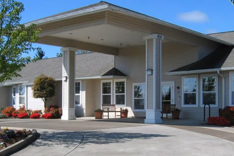 Exterior view of a single-story assisted living facility building with a covered entrance supported by two columns. There are benches and potted plants near the entrance, with landscaping including bushes and flowers around the building. The sky is clear and blue.